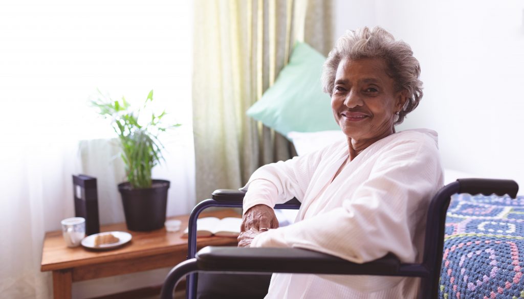 Portrait of happy mixed race senior woman sitting in wheelchair while looking at camera at nursing home